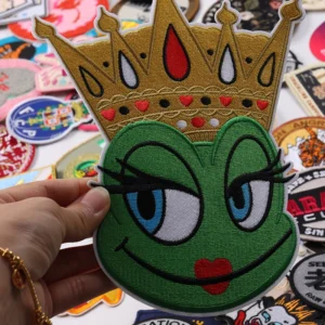 Green cartoon mascot face wearing a gold jeweled crown embroidered patch