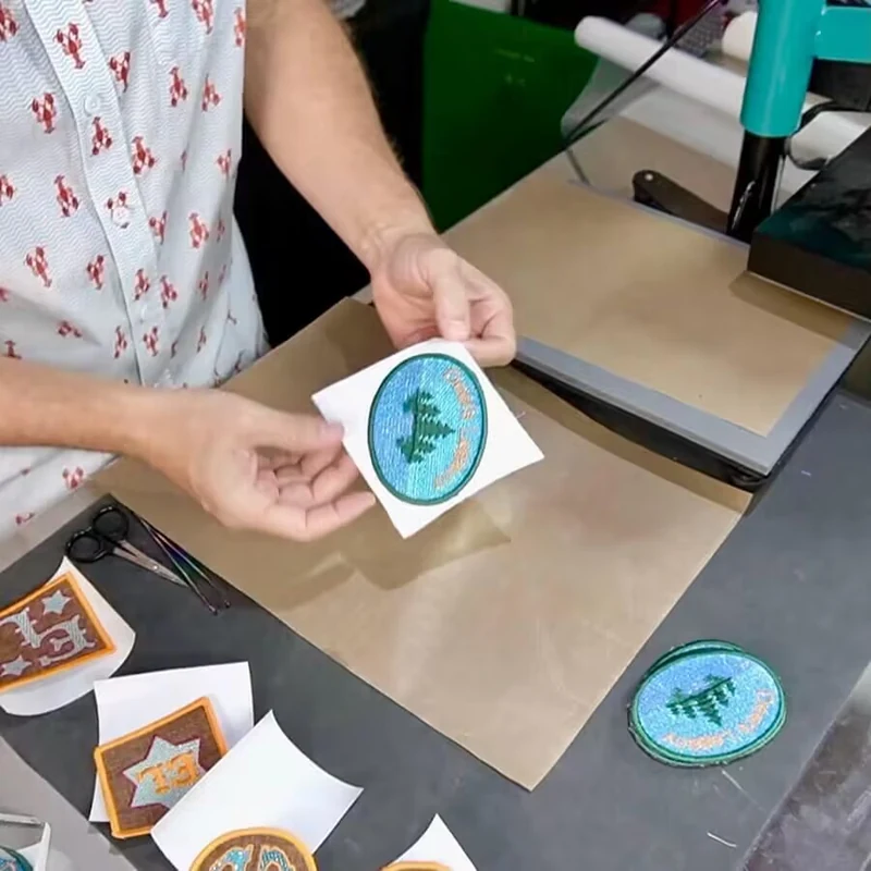 Person holding a small blue green patch beside a heat press workstation.
