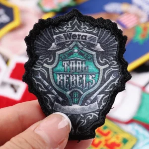 Black ornate embroidered badge with gray teal and white lettering held between fingers