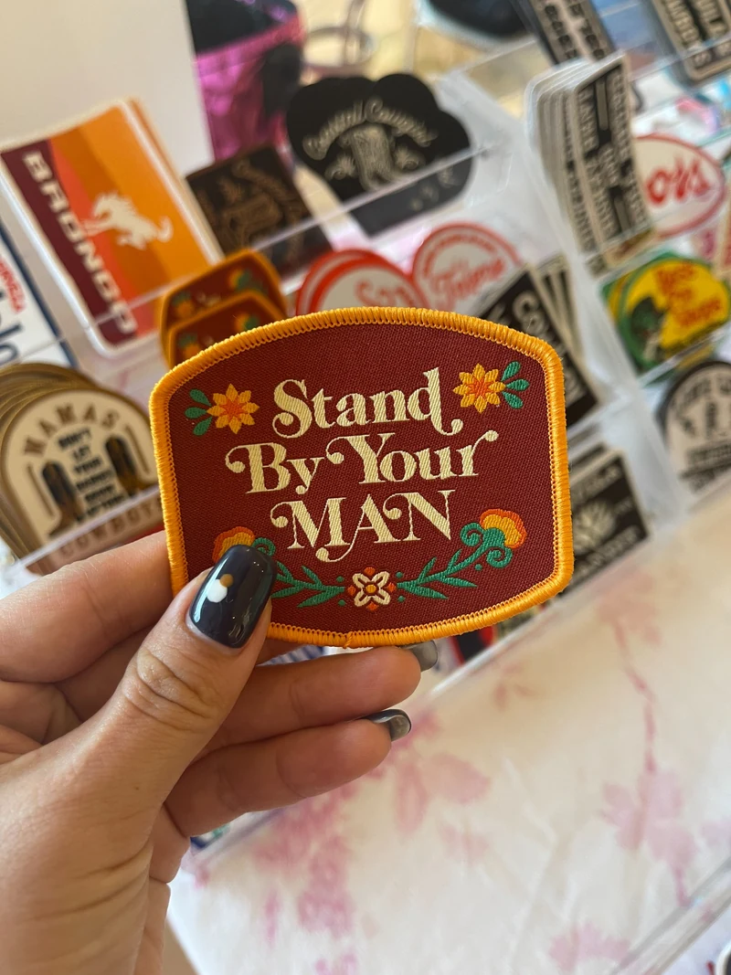 Hand holding a burgundy Stand By Your Man embroidered patch with yellow border and floral details.