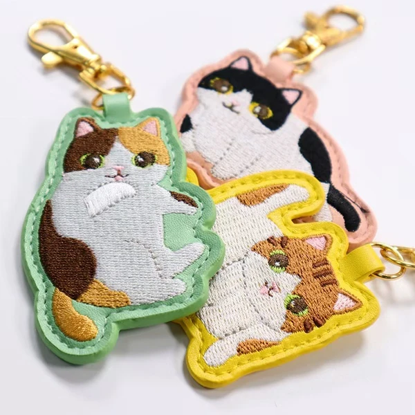 Three cat-shaped embroidered keychains with mint green, pink and yellow borders and gold-tone clasps.
