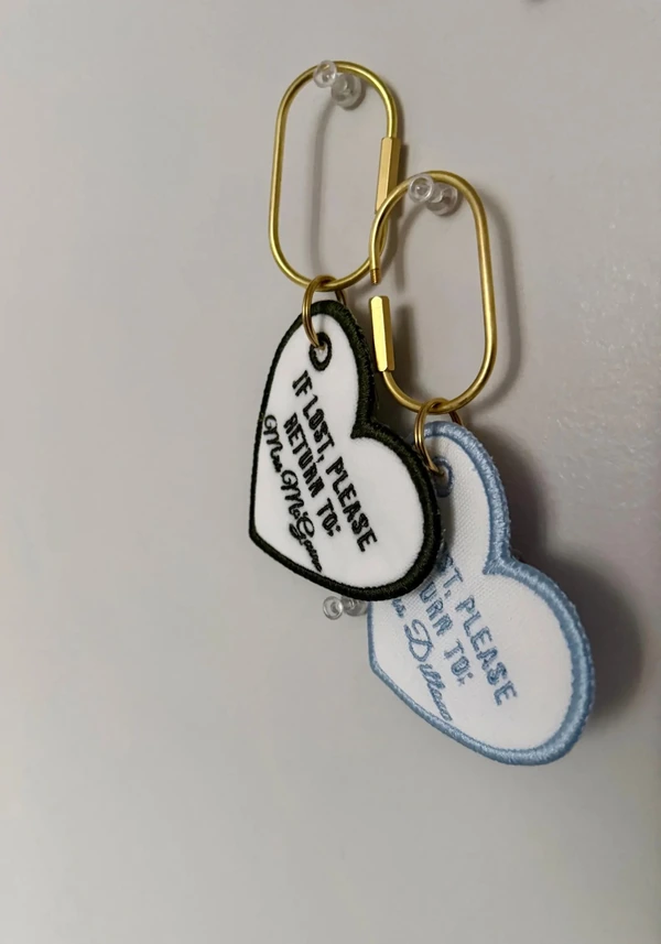 Two white heart-shaped embroidered keychains with black and light blue borders and gold-tone oval clips.