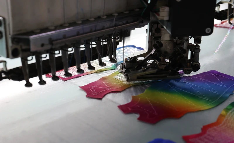 Close-up of an embroidery machine needle and presser assembly working over a pink yellow green blue and purple sequin panel.