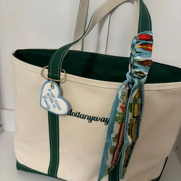 White heart-shaped bag charm with light blue embroidery attached to a cream and green canvas tote beside a patterned scarf.