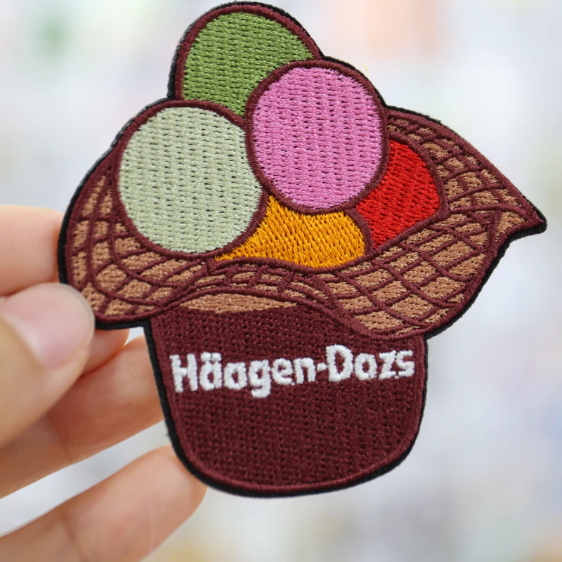 Hand holding a shaped ice cream basket patch with multicolored scoops brown basket stitching and white Haagen-Dazs lettering.