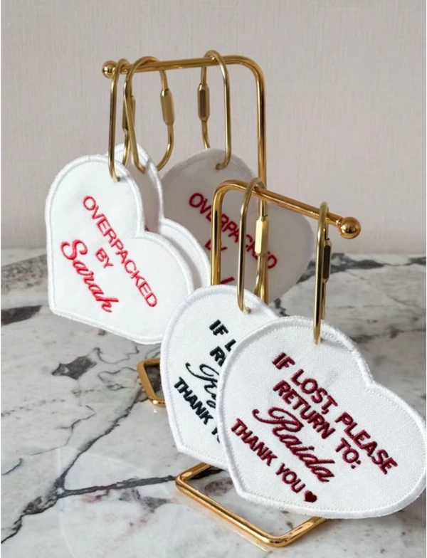 White heart-shaped fabric keychains with red, burgundy and black embroidered lettering displayed on gold-tone stands.