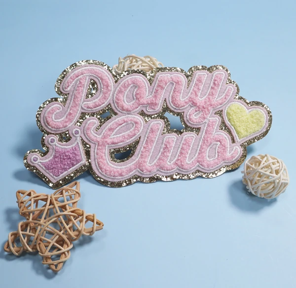 Pink Pony Club script chenille patch with a purple crown, yellow heart and sparkling border on a blue background with wicker decorations.