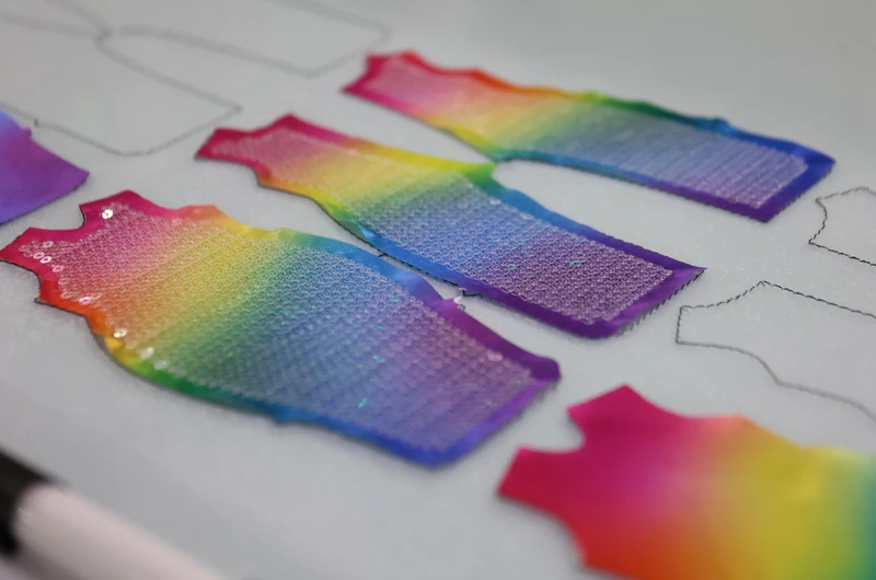 Close-up of vest-shaped rainbow sequin panels on white fabric, with sparkling rows and unfilled stitched outlines nearby.