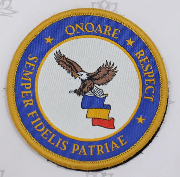 Round embroidered badge with an eagle above blue, yellow and red artwork, a blue text ring and yellow stitched border.
