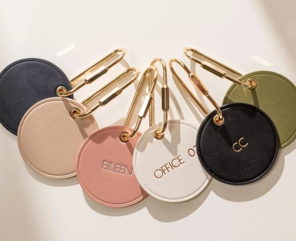 Six round leather-look bag charms in navy, beige, pink, white, black and olive with gold-tone oval clips; several show personalized lettering.