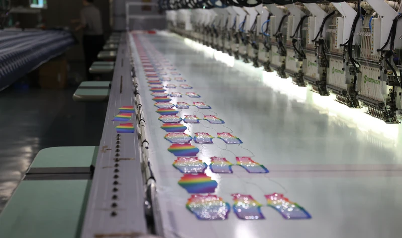 Wide view along a long row of embroidery machine heads with rainbow panels arranged on an illuminated white fabric bed.