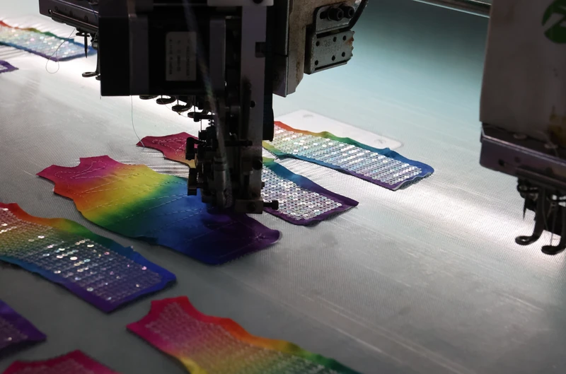 Angled view of a machine needle assembly above a rainbow vest-shaped panel, with other sparkling panels arranged on white fabric.