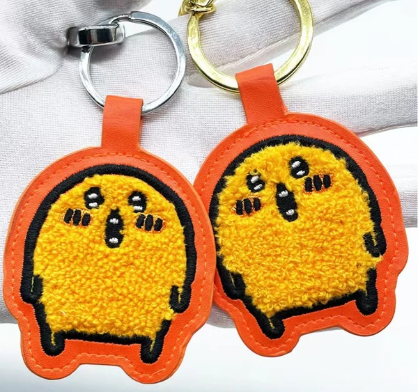 Two yellow character keychains with fluffy chenille texture, black embroidered details, orange borders and metal key rings.